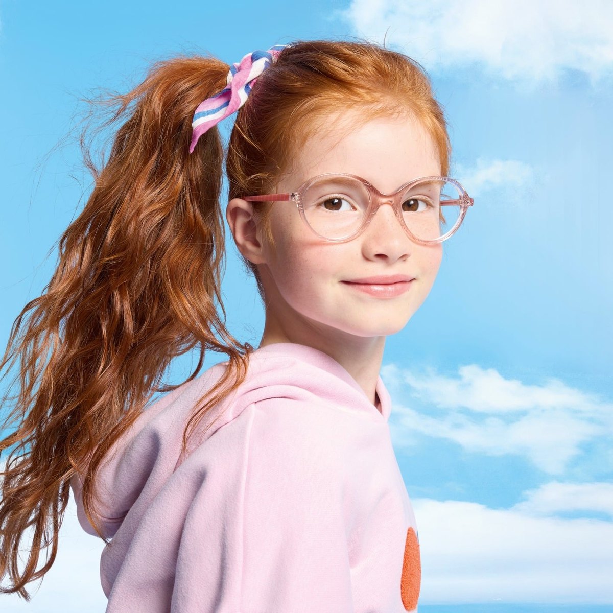 Fillette rousse au sourire discret, portant une monture Catimini transparente rose, sur fond de ciel bleu.