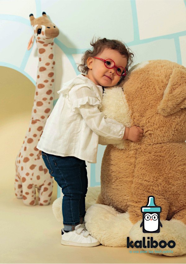 Bébé fille portant une monture Kaliboo rouge, blottie contre une peluche beige, devant un mur pastel.