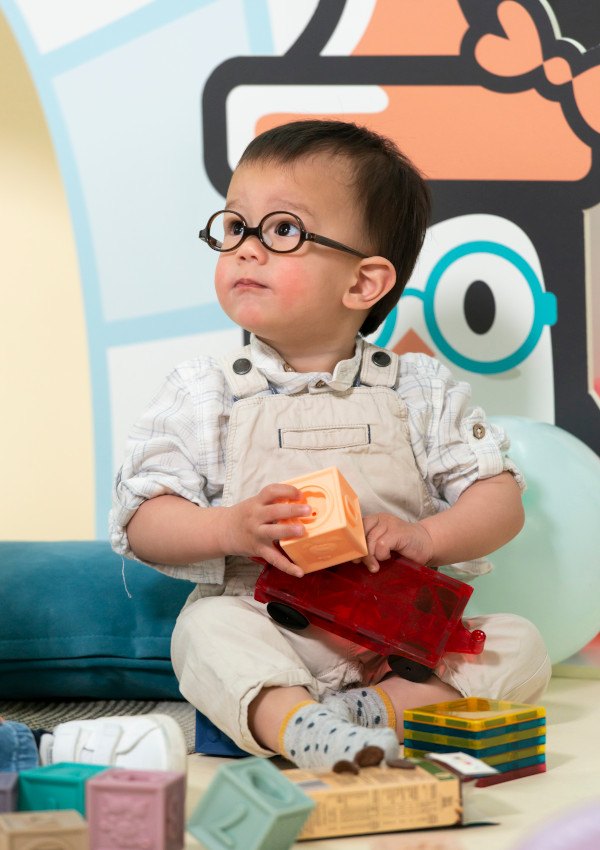 Bébé garçon portant une monture Kaliboo ronde marron, assis parmi des jouets devant un mur pastel.