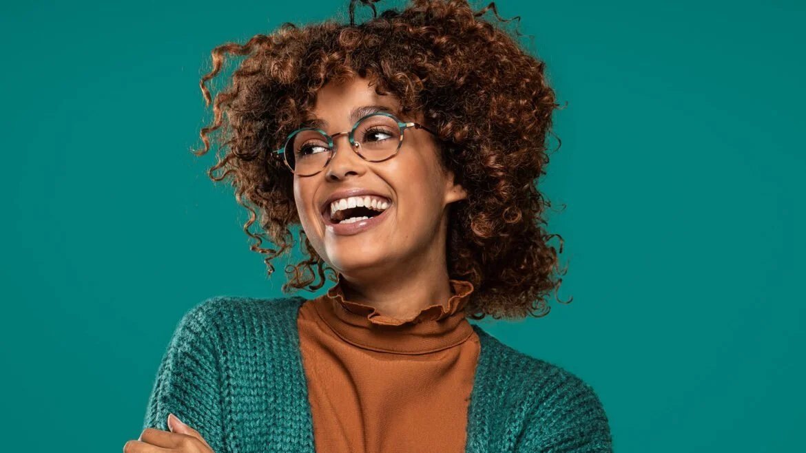 Femme souriante aux cheveux bouclés, portant une monture Nifties ronde, sur fond turquoise.
