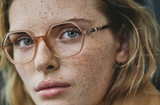 Gros plan sur le visage d'une femme aux taches de rousseur, portant une monture Nifties hexagonale transparente.