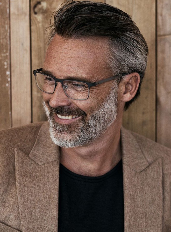 Portrait d'un homme souriant, portant des montures Prodesign sombres à fines lignes, devant un mur en bois.